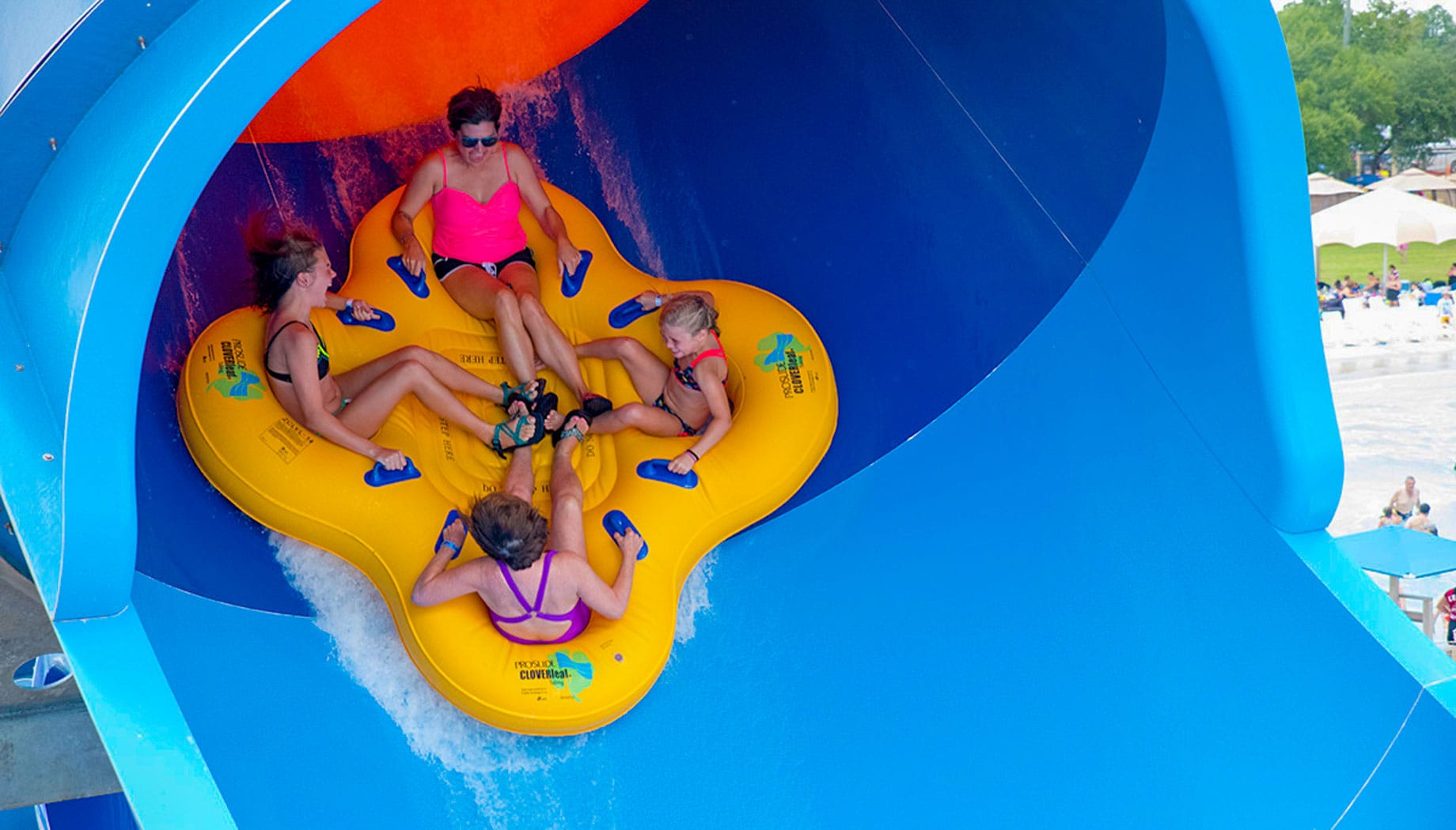 rays-riptide-family Four people are riding a bright yellow inflatable raft down a blue water slide at a water park.