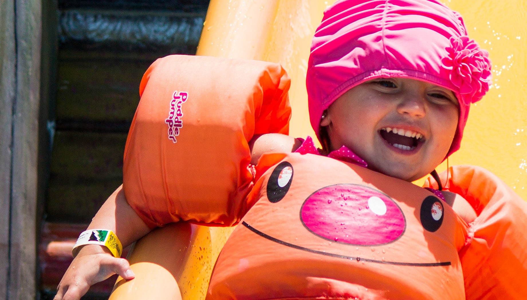 toddler-waterpark A young child in orange floaties and a pink swim cap smiles while sliding down a water slide.